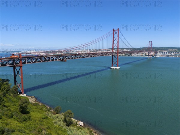A large red bridge stretches across a wide expanse of water under a clear blue sky, surrounded by