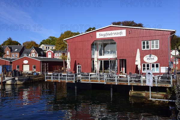 A red wooden building with 'Hotel' lettering on the waterfront under a clear blue sky on a sunny