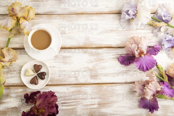 Cup of cioffee with chocolate candies, lilac and purple iris flowers on white wooden background.