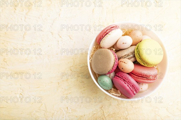 Multicolored macaroons and chocolate eggs in ceramic bowl on beige concrete background. top view,