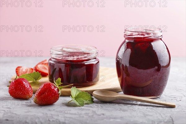Strawberry jam in a glass jar with berries and leaves on gray concrete background. Homemade, close