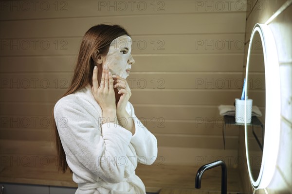 Woman applying sheet mask with mineral elements on her face in front of a mirror