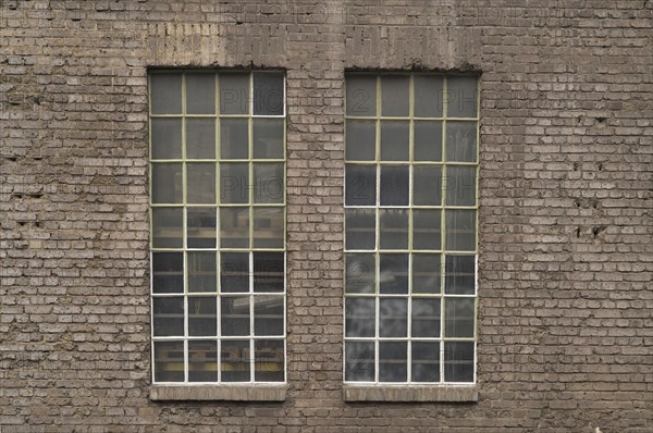 Two large industrial windows with small panes and metal frames are set in an old brick wall,