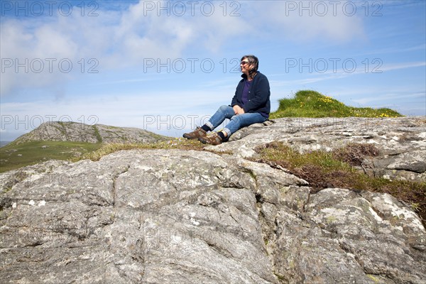 Middle aged female hiker sitting high on rocky outcrop in Barra, Outer Hebrides, Scotland, UK
