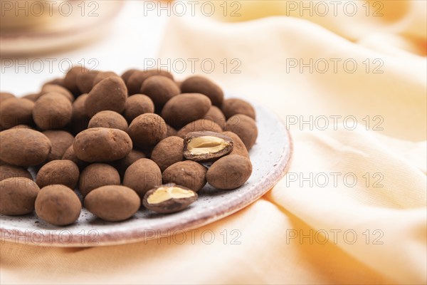Almond in chocolate dragees on ceramic plate and a cup of coffee on white concrete background and