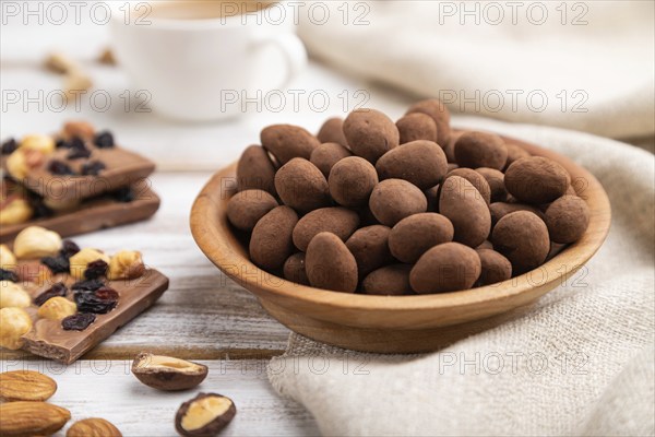 Almond in chocolate dragees in wooden plate and a cup of coffee on white wooden background and