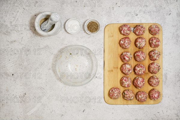 Top view of raw meatballs on wooden cutting board next to empty bowl and spices