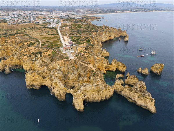 Steep cliffs and rocky coastline with blue sea, beyond a town and distant mountains, aerial view,