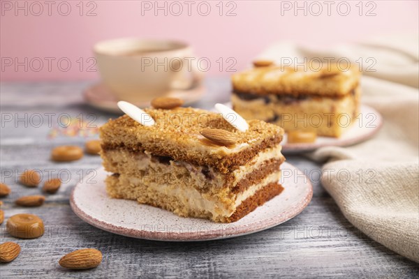 Honey cake with milk cream, caramel, almonds and a cup of coffee on a gray and pink background and