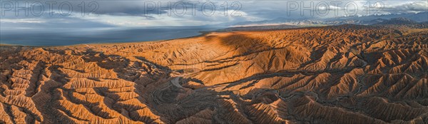 Panorama, landscape of eroded hills, badlands at sunset, behind Issyk Kul Lake and mountain peaks