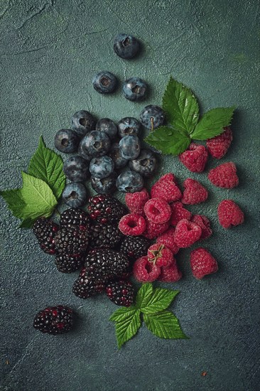 A mix of ripe berries, with leaves scattered on the table, raspberries, blackberries, blueberries,