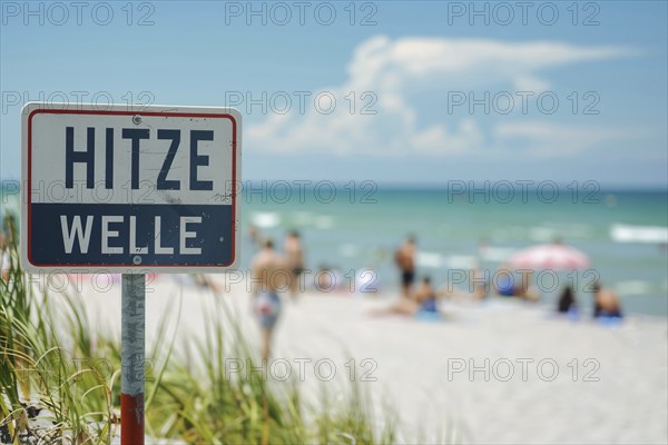 Warning sign with German text 'Hitzewelle' (Heat wave) in front of blurry sunny beach with people.