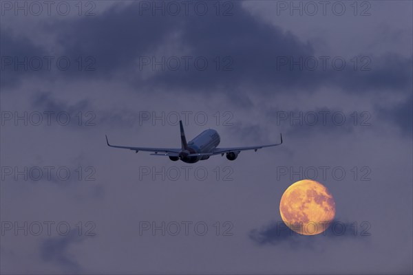 An aeroplane flies through the cloudy sky at night in front of a large, shining full moon, super