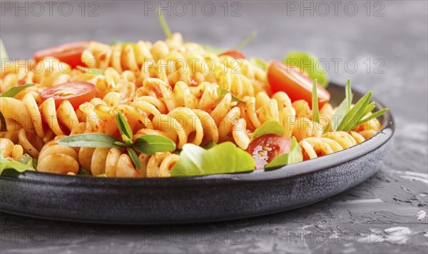 Fusilli pasta with tomato sauce, cherry tomatoes, lettuce and herbs on a black concrete background.
