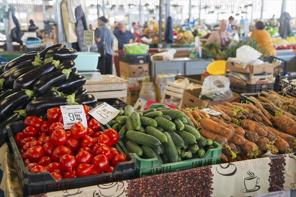Colourful display of vegetables at a market stall with price tags and background activity, market