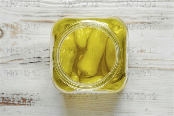 A glass jar of pickled peppers sits on a white wooden table. The jar is filled with whole, yellow