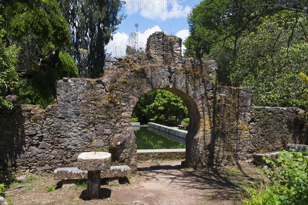 Ein alter Steinbogen führt in einen grünen Garten an einem sonnigen Tag, Mata Nacional do Buçaco,