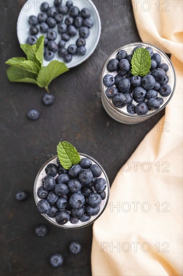 Yogurt with blueberry and chia in glass on black concrete background and orange linen textile. top