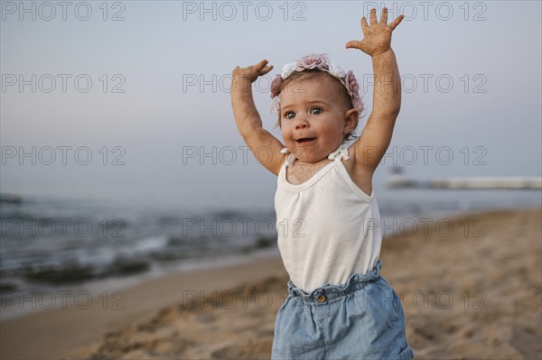 A baby in summery clothes and flowers in her hair stands on the beach at sunset and stretches both