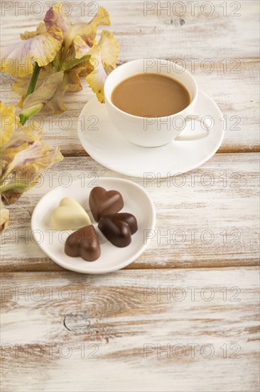 Cup of cioffee with chocolate candies, lilac and purple iris flowers on white wooden background.
