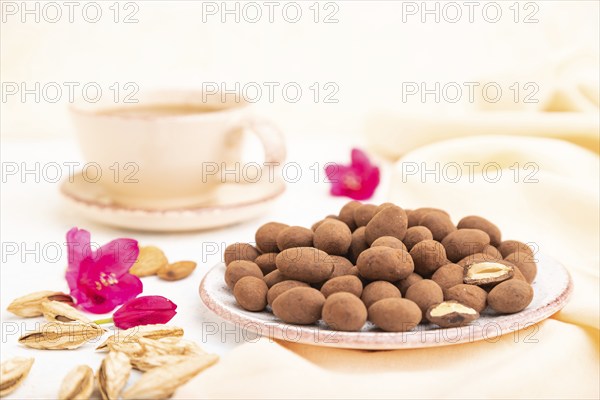 Almond in chocolate dragees on ceramic plate and a cup of coffee on white concrete background and