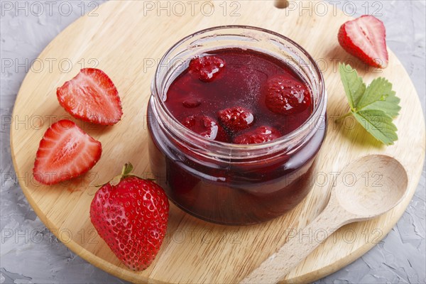 Strawberry jam in a glass jar with berries and leaves on gray concrete background. Homemade, close