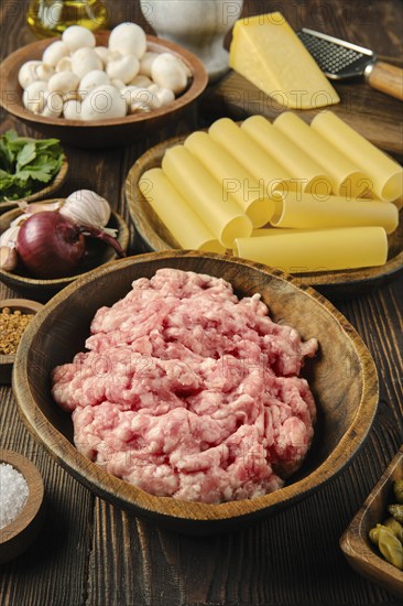 A wooden bowl filled with raw ground meat is surrounded by pasta, mushrooms, garlic, and cheese on