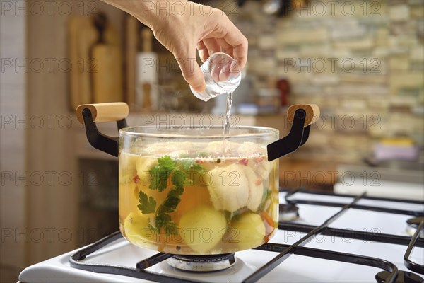 A hand adds vodka to a pot with fish soup, vegetables and herbs on a stove. The kitchen features a
