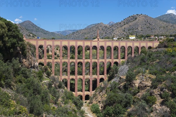 An old brick aqueduct spans a wooded ravine against a mountain backdrop under a clear blue sky,