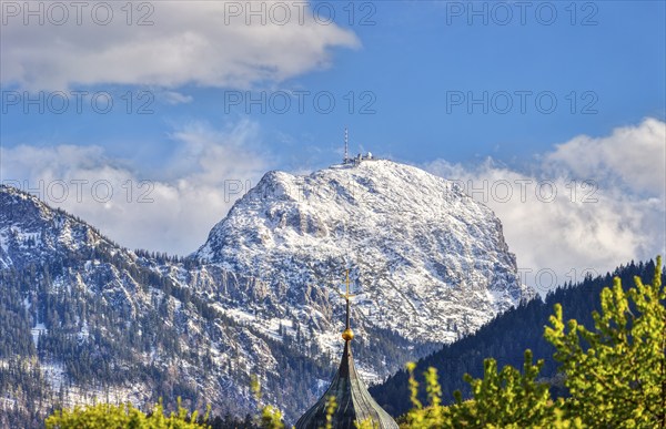 Snow-covered mountain peak under a blue sky with clouds, in the foreground a church tower and green