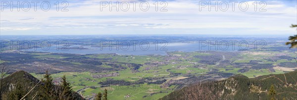 Panoramic view over a green landscape with mountains in the foreground and the large Chiemsee in