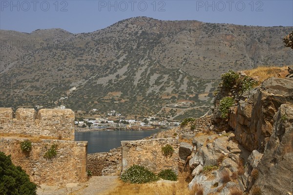 The remains of ancient stone structures in front of a mountainous landscape with a nearby village,