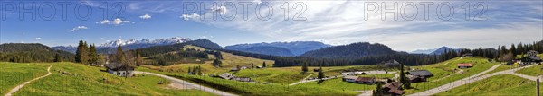 Wide green meadows in front of a mountain range, dotted with huts, under a partly cloudy blue sky,