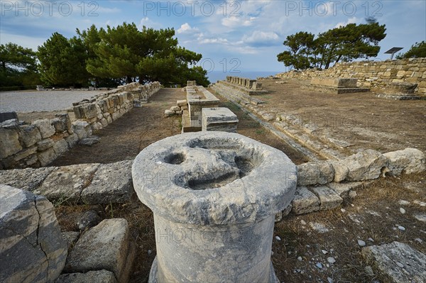 Ancient stone column with eroded details, surrounded by ruins and trees, Exedra, Kamiros,