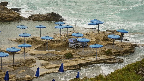 Rocks on the coast with blue parasols and deckchairs, surrounded by the sea with wavy water, Oasis