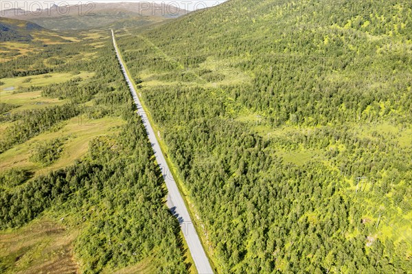 Aerial view of a straight road and power line cutting through forest and meadows on Senja island,