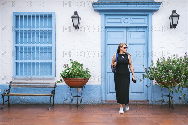 A fashionable woman in a black dress and sneakers walks past a blue colonial-style door and window