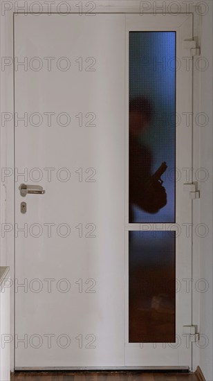 A burglar stands in front of an entrance door in the dark with a gun and looks through the glass