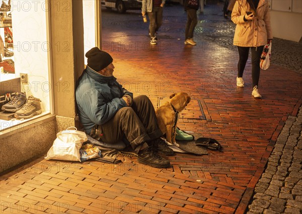 Homeless man with a dog sits in a shopping mall as people walk by in Husum, Schleswig-Holstein,