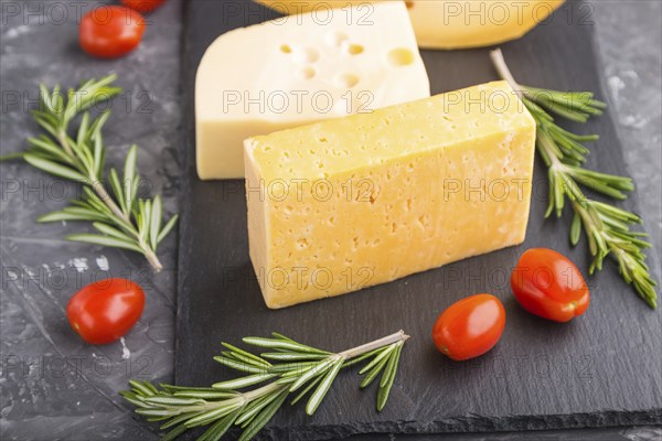 Various types of cheese with rosemary and tomatoes on black slate board on a black concrete