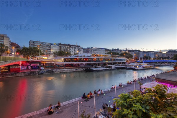 Evening scene by the river with illuminated promenade and people on the riverbank, relaxed