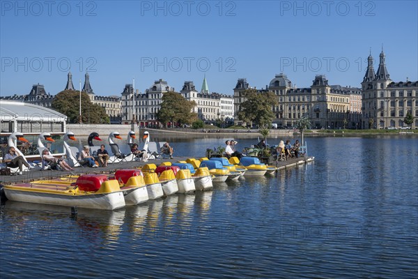 People enjoying leisure time on a jetty by the water, pedal boats, blue sky, historic buildings,