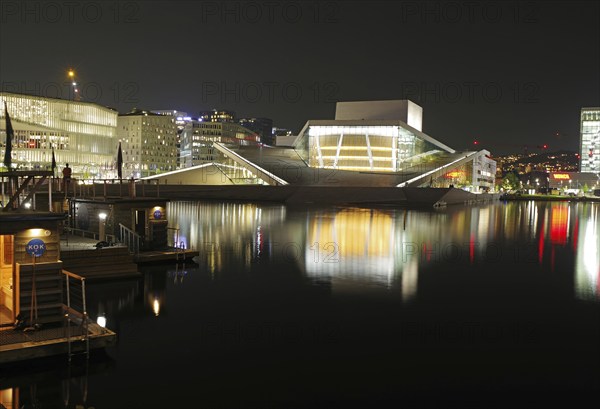 Illuminated opera house and modern buildings reflected in calm waters at night, Oslo city centre,