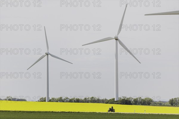 A motorbike on a country road stands out in front of a rapeseed field and wind turbines in