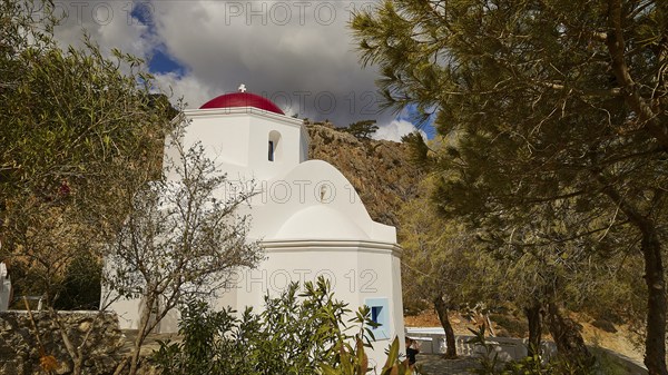 A picturesque white church with a red dome, surrounded by trees and rocks under a changeable sky,