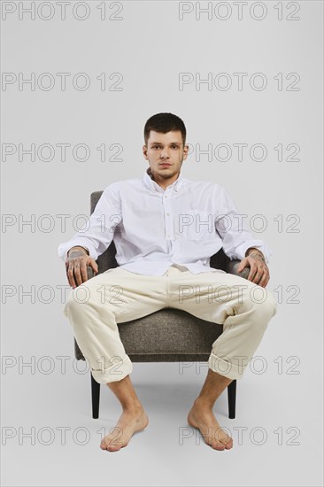 Full length studio portrait of young barefoot man in white shirt and ivory trousers sitting in