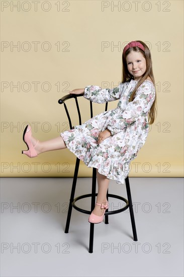 Cute little girl in sundress and hair band sits on tall chair in studio over pale yellow background