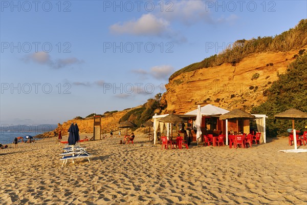 Beach café, red rocks illuminated by the evening sun, sandy beach with tourists, holiday by the