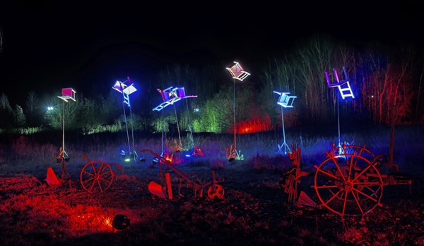Artwork with colourful lights and chairs above wagon wheels at night, surrounded by trees, world of