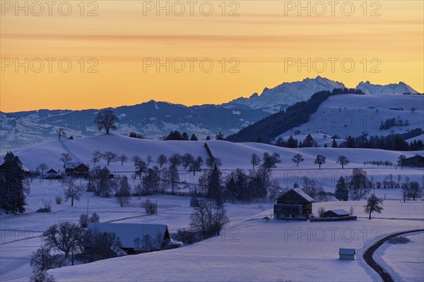Snowy landscape at sunrise with mountains and farms, bathed in a warm orange-blue light, Menzingen,